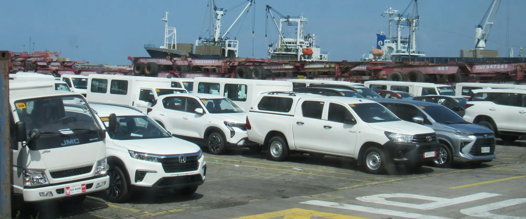 Cars lined up on a port apron, with cargo ships and a crane behind them.